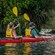 Two Guests with life jackets paddling a double kayak along a forested shore
